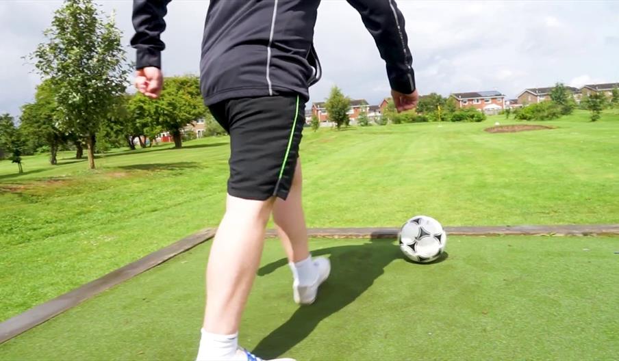 A person in shorts goes to kick a football on a wide green patch with fields dotted around and a small flag in the distance.