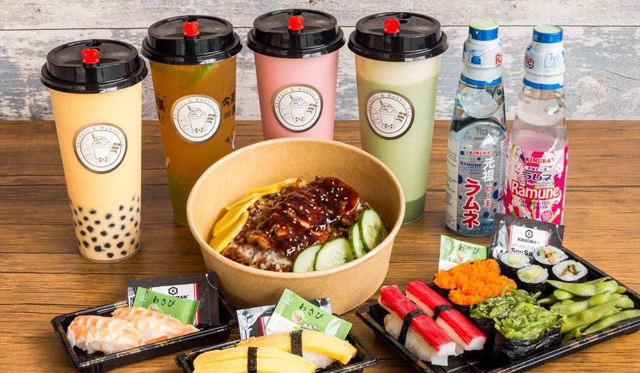 A variety of Japanese foods and drinks on a wooden table, including sushi, nigiri, a rice bowl topped with grilled meat and cucumbers, bubble tea drin