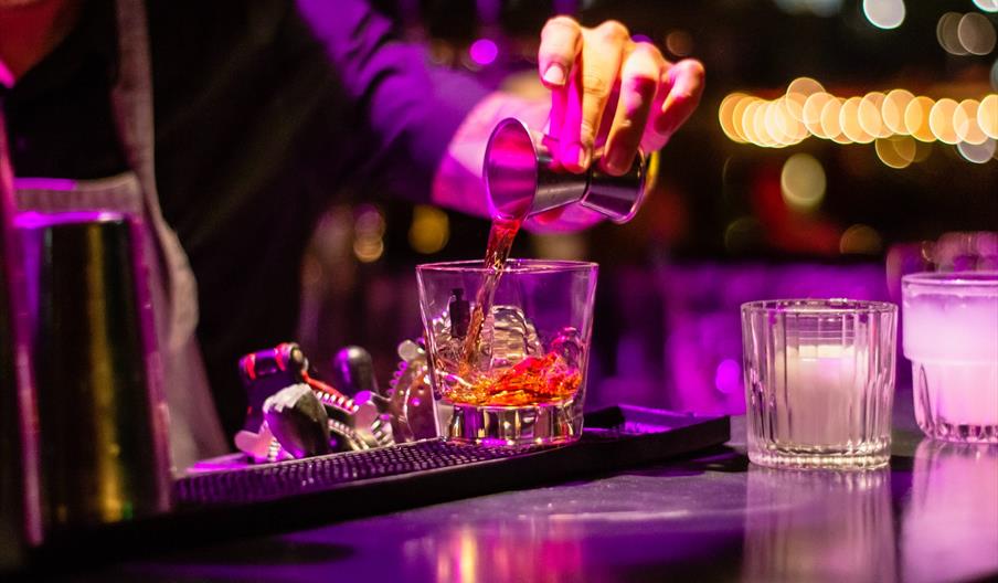 Interior of The Star. A bartender, illuminated by purple and yellow lights, pours a brown liquor from a jigger into a glass with ice on a bar counter,