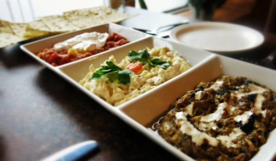 A white rectangular dish is divided into three sections, each filled with a different dip or spread, garnished with vegetables and herbs. In the backg