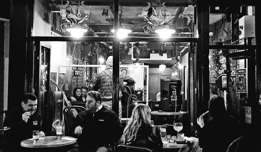 Black and white photo of people sitting at tables outside Mr. Moody's Tavern at night, with others visible inside and a musician playing near the bar'