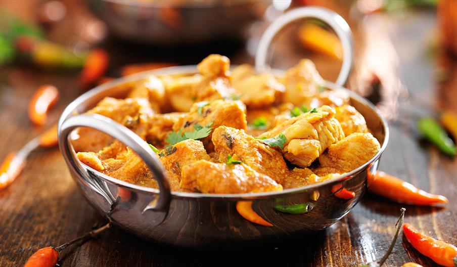 A metal bowl filled with pieces of chicken curry garnished with fresh cilantro, with whole red and green chili peppers scattered on the wooden table.
