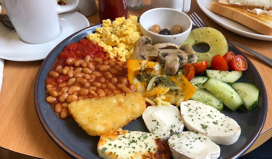 A breakfast plate with baked beans, scrambled eggs, grilled tomatoes, sautéed mushrooms, hash brown, pineapple ring, cherry tomatoes, cucumber slices,