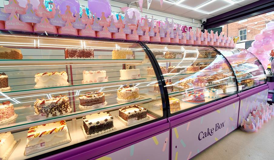 A row of glass cabinets displaying a selection of celebration cakes along with  pink ballons and cardboard crowns.