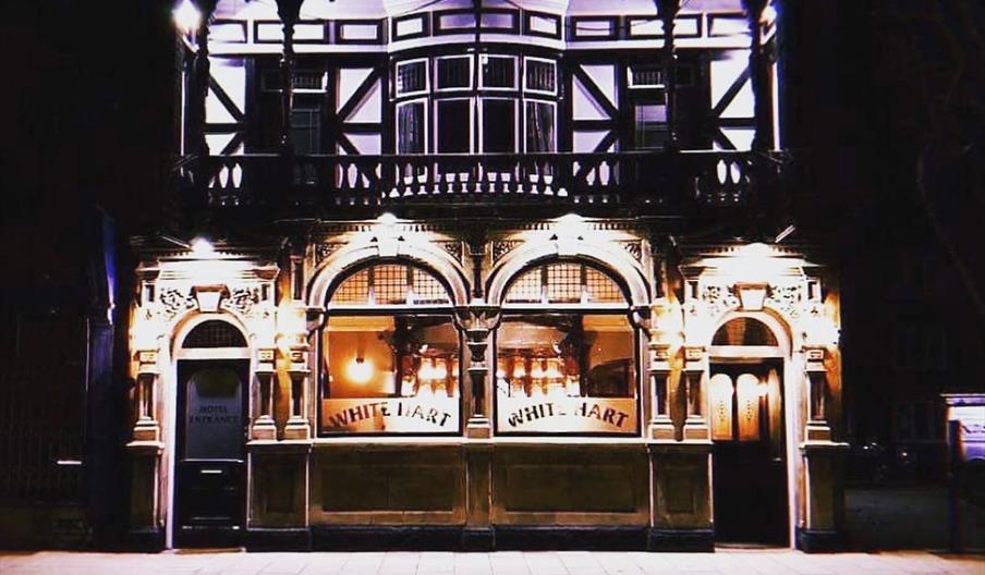 A brightly lit pub with ornate architecture and large arched windows, featuring the name "White Hart" displayed in both front windows. The upper floor