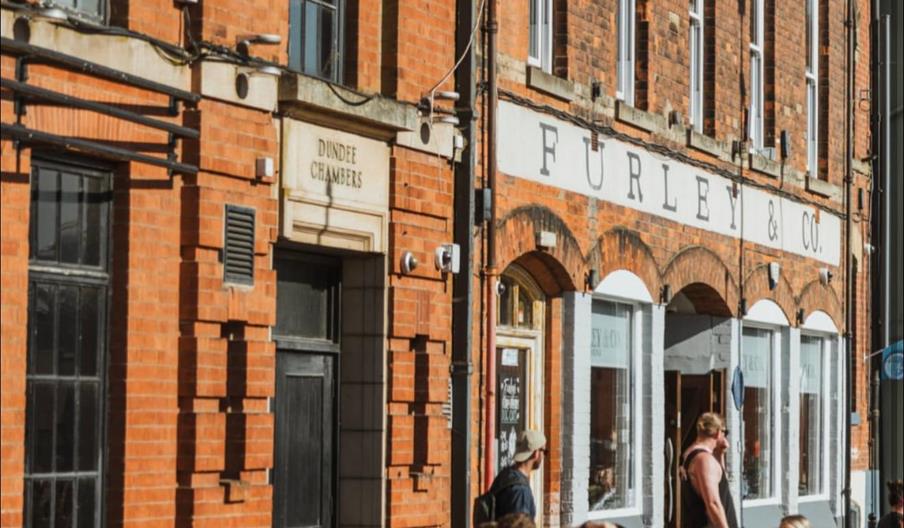 People sit at outdoor tables enjoying drinks and conversation in front of a red brick building with sign reading and "Furley & Co." on a sunny day.