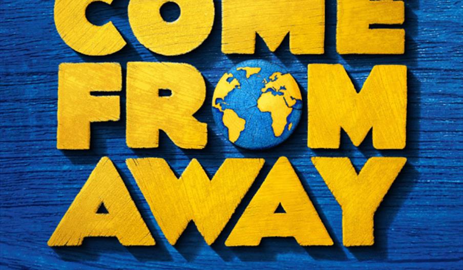 The image is a promotional poster for the musical "Come From Away," featuring large, bold yellow letters on a blue wooden background, with a depiction