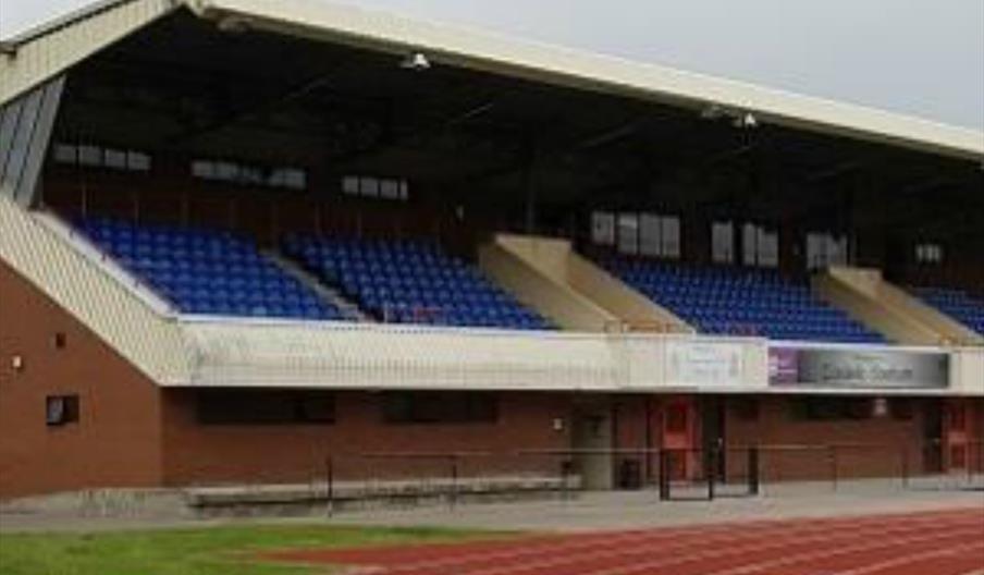 A covered Costello Stadium seating area with rows of blue seats overlooks a red running track under a cloudy sky.