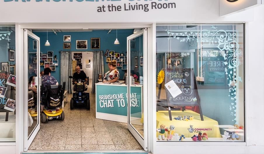 Entrance to a community space called Bransholme Chat at the Living Room, with open glass doors, people inside talking, and a welcoming reception area