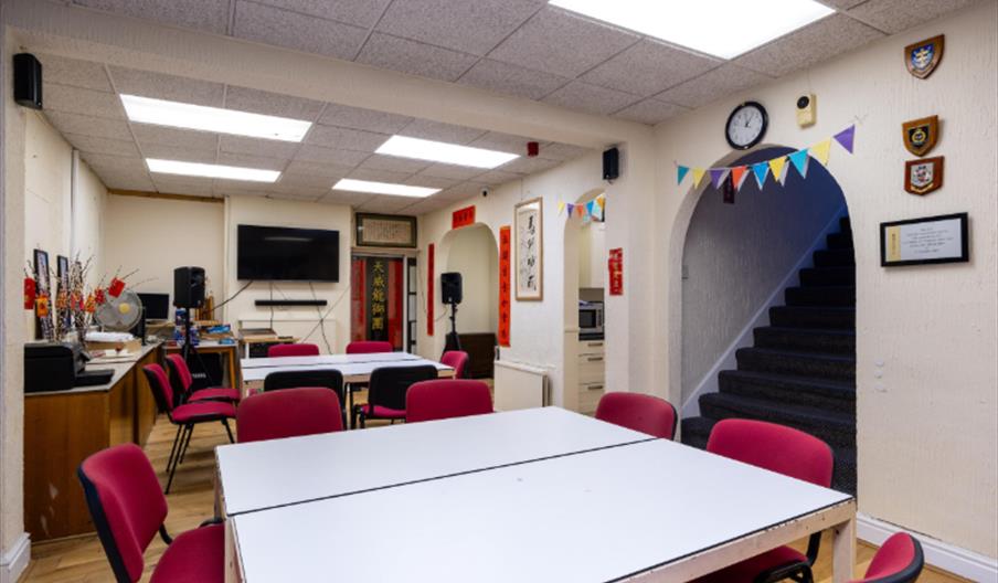 A brightly lit meeting or classroom with white tables and red chairs arranged in groups, decorated with colorful bunting, Chinese scrolls, plaques on