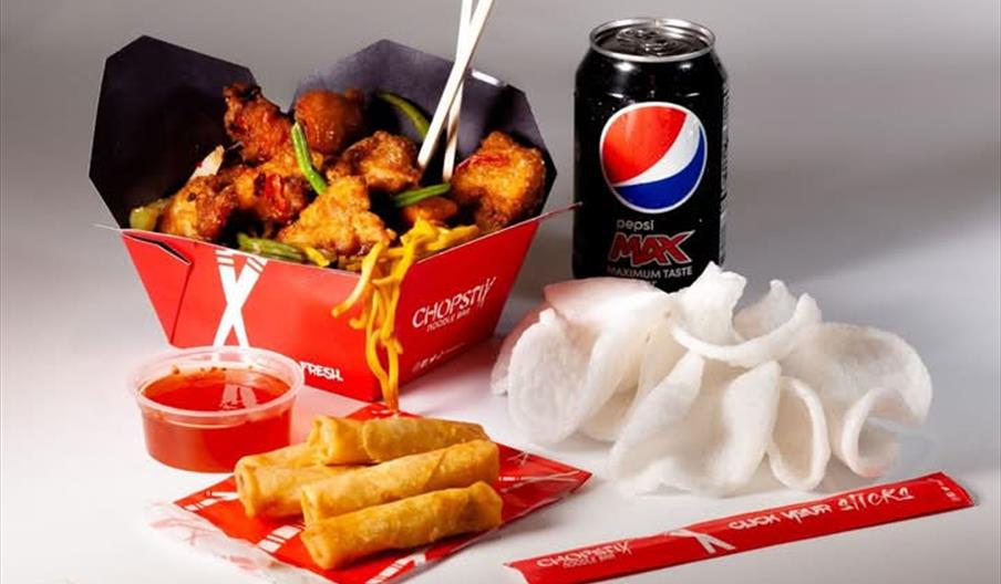 A takeout meal featuring a box of stir-fried noodles with vegetables and chicken, a can of Pepsi Max, a serving of prawn crackers, two spring rolls wi