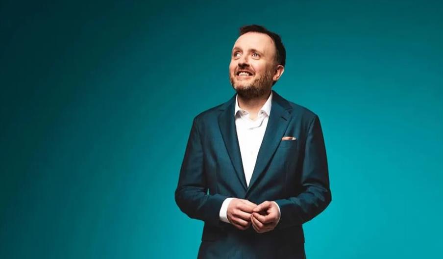 Chris McCausland stands in a dark blue suit and white shirt against a teal background, looking upward and smiling with his hands loosely clasped in fr