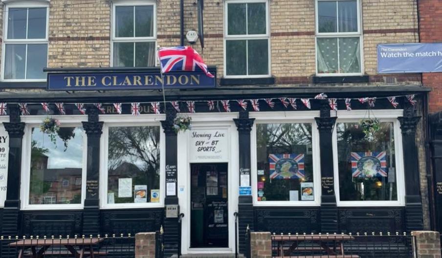 Front view of The Clarendon pub, a two-story brick building with a Union Jack flag above the entrance, patriotic bunting, and benches outside. Large w