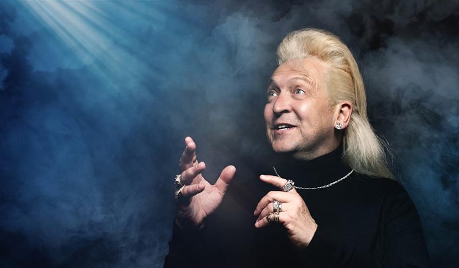 Clinton Baptiste dressed in a turtleneck with jewelry on poses in a dark smoky room.