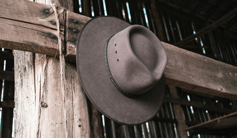 A brown cowboy hat hangs on a wooden beam inside a rustic, weathered barn.
