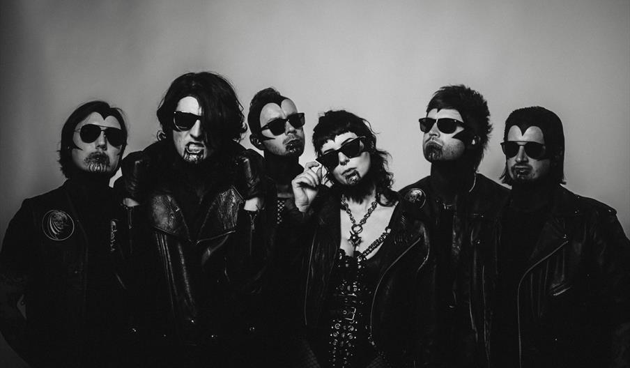 Six people wearing dark sunglasses, black leather jackets, and gothic makeup stand together against a plain background in a black and white photo, giv