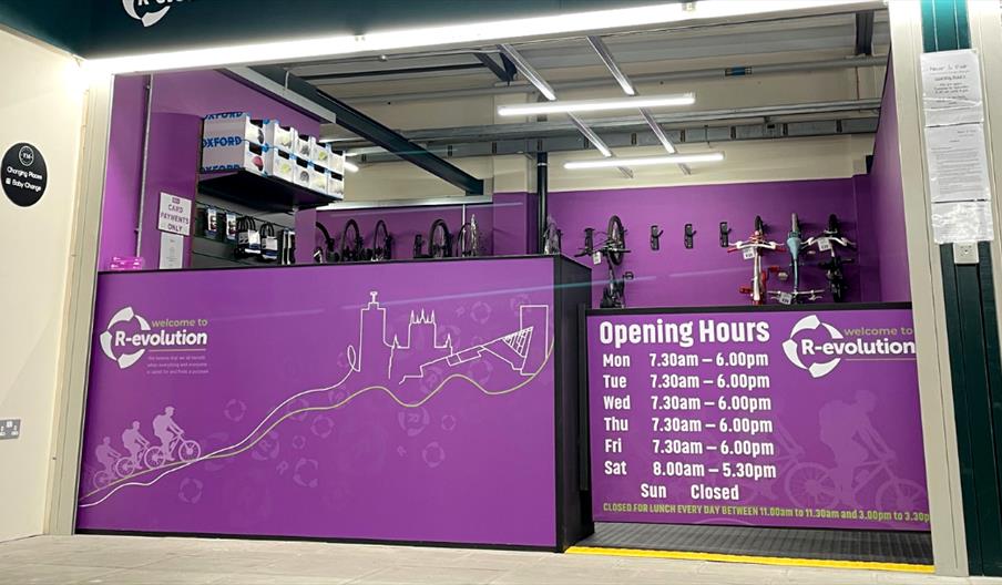 The Cycle Hub with purple walls and a front counter displaying the R-evolution logo, opening hours, and a cityscape line drawing; bicycles and bike ac