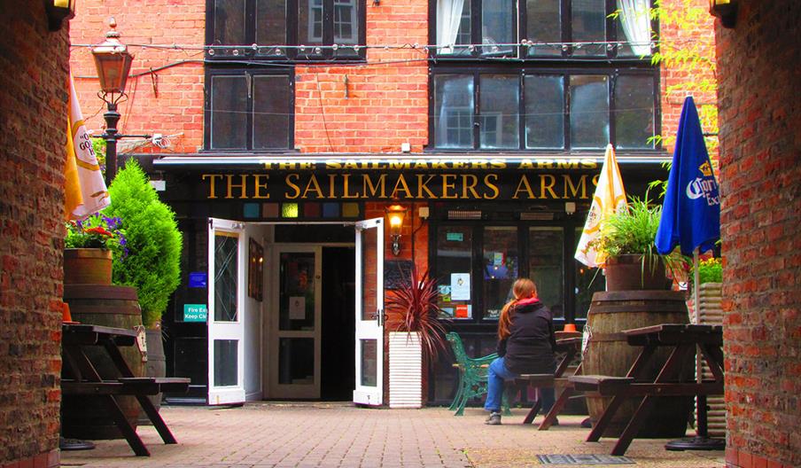 View of the entrance to "The Sailmakers Arms," with brick walls, open double doors, outdoor wooden tables, large plant pots, and a person sitting alon