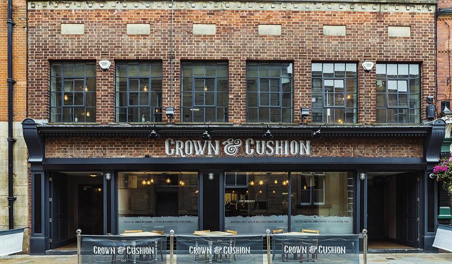 Exterior of red brick building, windows with grey trim, Crown and Cushion painted on the wall and metal table and chairs.