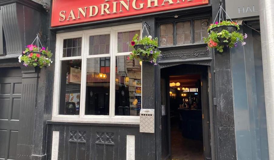 The entrance to the pub "The Sandringham" with a large red sign above the door and window, two hanging flower baskets on either side of the entrance,