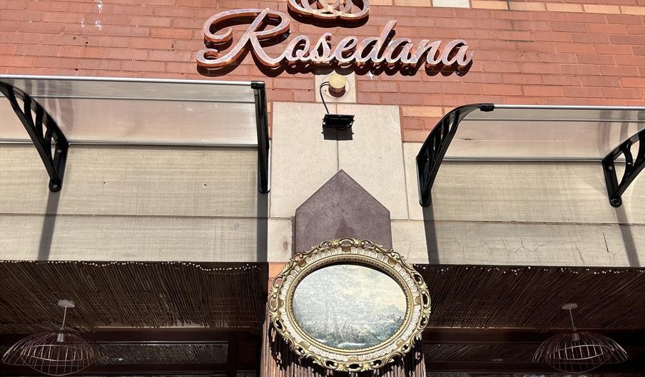 Signage for "Rosedana" restaurant, featuring a decorative emblem and framed vintage artwork against a brick wall with floral curtains.