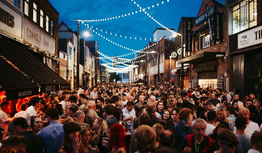 A large crowd of people fill Humber Street, with several bars and eateries on either side and lit fairy lights weaving their way between lampposts abo