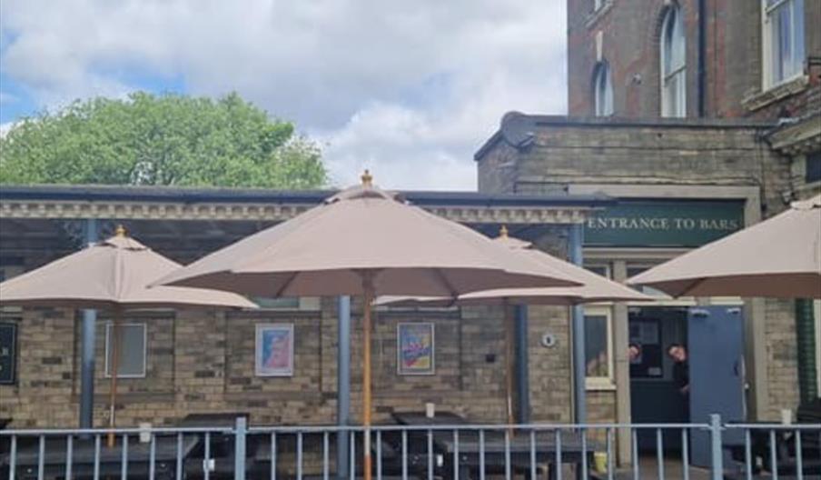 Outdoor seating area of The Halfway Hotel with dark wooden picnic tables and large beige umbrellas, enclosed by a metal fence, in front of a brick bui
