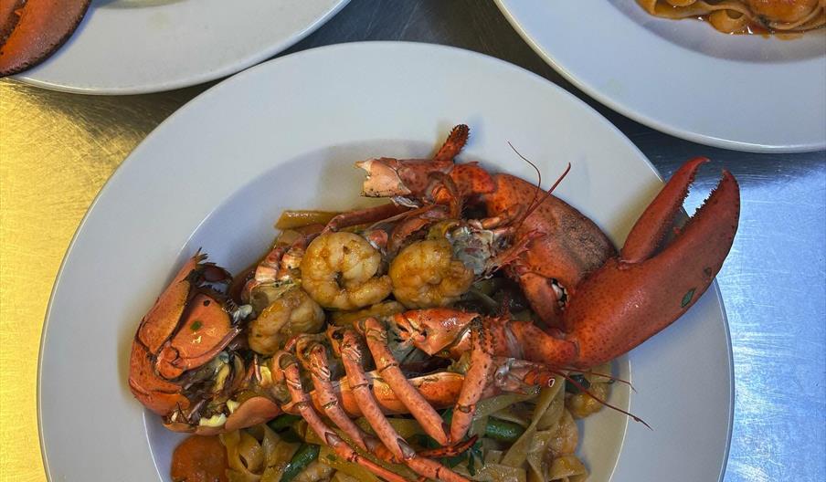 A close-up view of three plates of seafood pasta dishes, with the central plate featuring a whole cooked lobster and shrimp atop fettuccine with veget