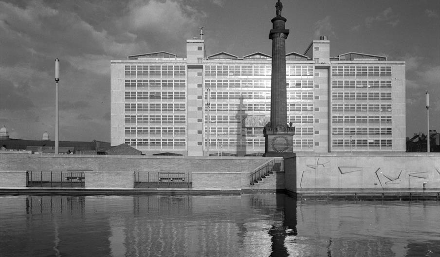 A greyscale picture showing a statue on top of a tall pillar standing in front of a large modern looking building, whilst in front of the statue a rai