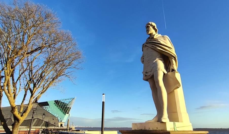 A Statue of a man in front of a tree with no leaves and the exterior of The Deep, an angular modern building, each backed by a blue sky.