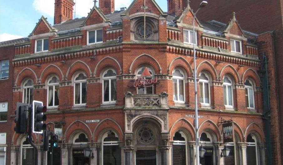 A historic red-brick Victorian building on a street corner houses The Dram Shop, with arched windows, decorative stonework, and several chimneys, as p