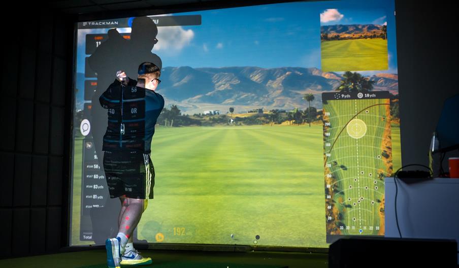 A young man in shorts swings a golf club on a small turf, with a projection of a golf course in front of him.
