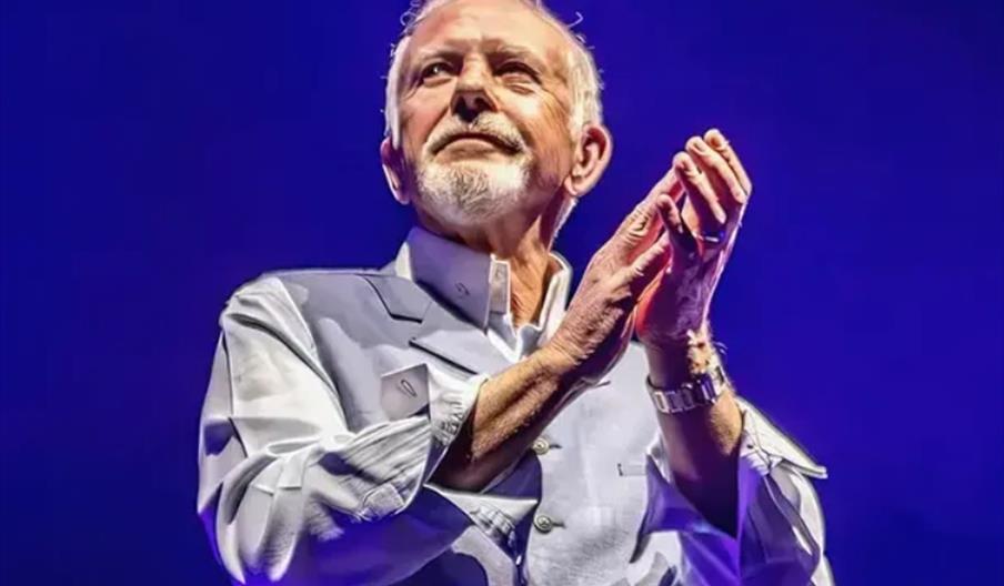 David Essex stands under dramatic lighting against a blue background, wearing a light-colored shirt and vest, with his hands raised as if applauding o