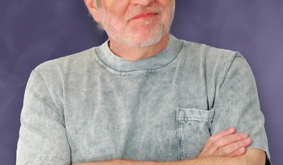 Older man with gray hair and beard standing with arms crossed, wearing a light gray t-shirt, against a purple marbled background.