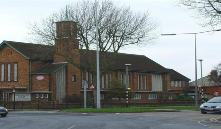 A brick church with a tall central tower and large vertical windows sits on a street corner, surrounded by bare trees, cars, and nearby residential ho