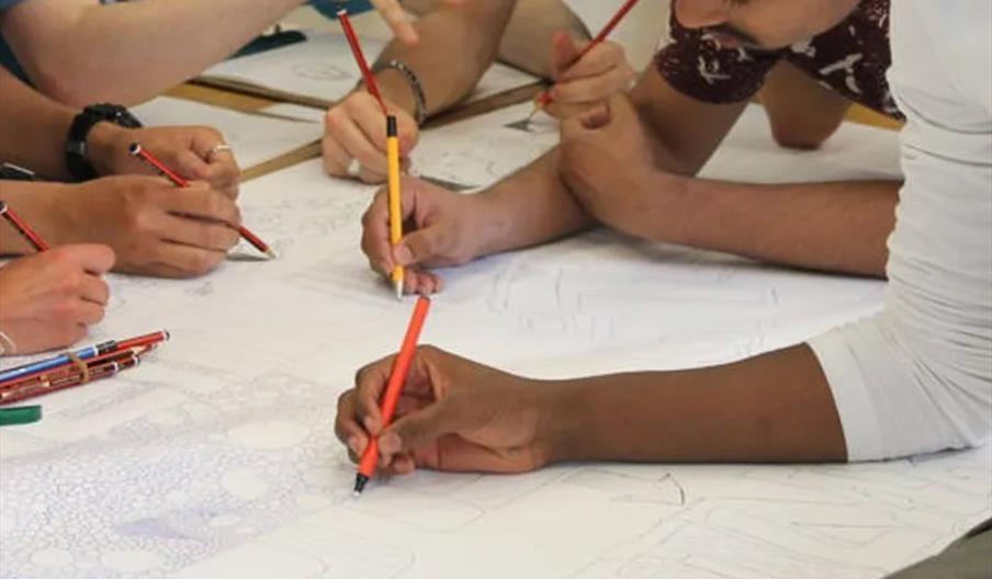Several people are gathered around a large sheet of paper, each holding colored pencils and collaboratively drawing or coloring on the paper.