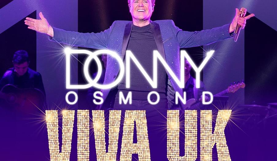A man wearing a sparkly blue jacket and holding a microphone stands on stage with his arms open wide, smiling. Bold, sparkling text across the image r