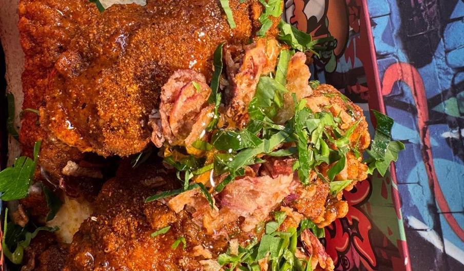 A serving of crispy fried chicken coated with spices, garnished with chopped herbs and thin fried onions, placed on a slice of bread in a colourful gr