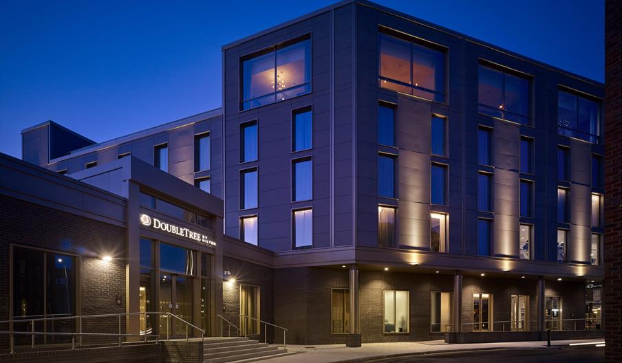 A modern, multi-story DoubleTree by Hilton hotel building is lit up at dusk, with large windows and a well-lit entrance featuring a sign above the doo