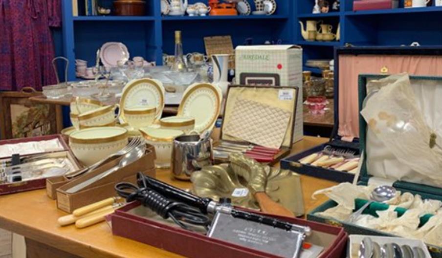 A wooden table is covered with assorted vintage items, including cutlery sets, plates, boxed utensils, and small household goods, while shelves in the