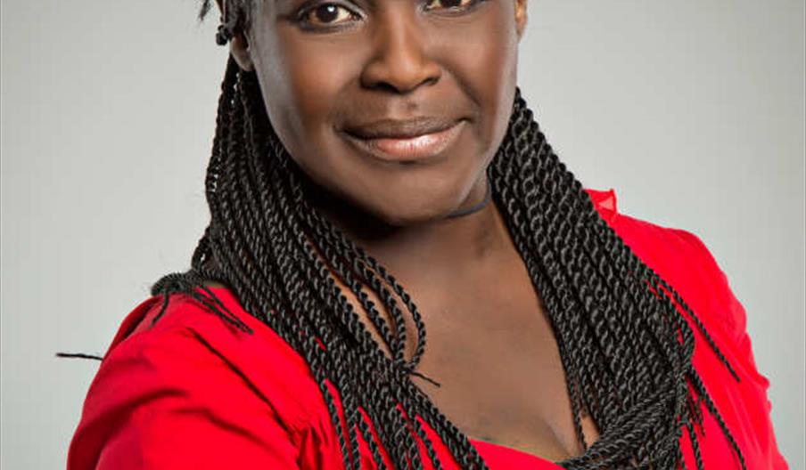 Dame Maggie Aderin-Pocock is wearing a bright red top and smiling slightly while posing against a plain gray background.