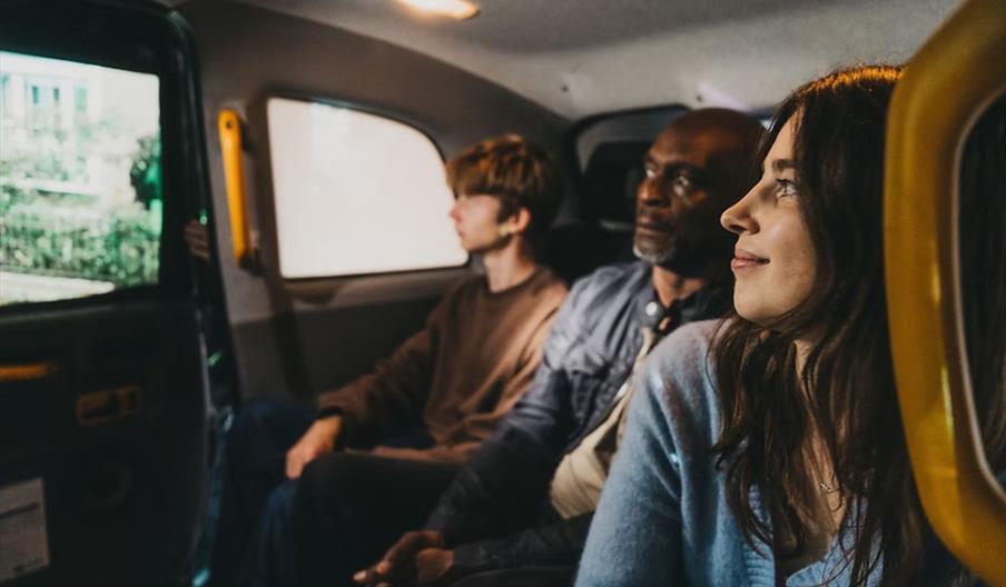 Three people are sitting in the backseat of a vehicle, looking out the windows; the person closest to the camera is smiling, and the vehicle door is o