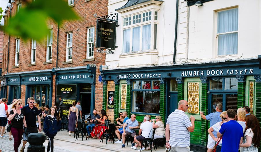 Groups of people are walking and sitting outside the Humber Dock Bar, a bar and grill with blue and green trim, on a lively street. Some people are ch