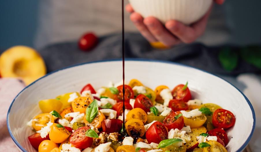 A person is drizzling dark balsamic dressing from a spoon over a bowl of colorful cherry tomatoes, mozzarella, and basil salad. Fresh tomatoes and bas