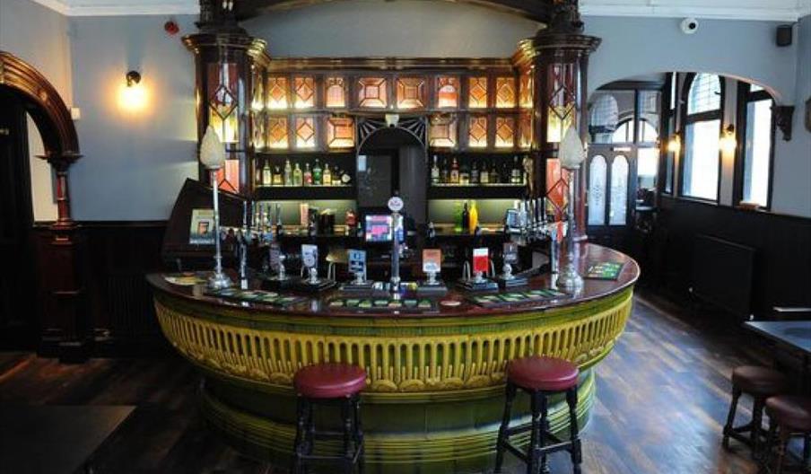 An ornate, vintage-style bar with a wooden counter and decorative yellow front, set in a warmly lit pub interior with wooden floors, tall bar stools,
