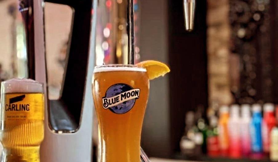 Interior of The Bottle Top bar. A tall glass of Blue Moon beer with a slice of orange on the rim sits on a bar counter in front of a Blue Moon draft t