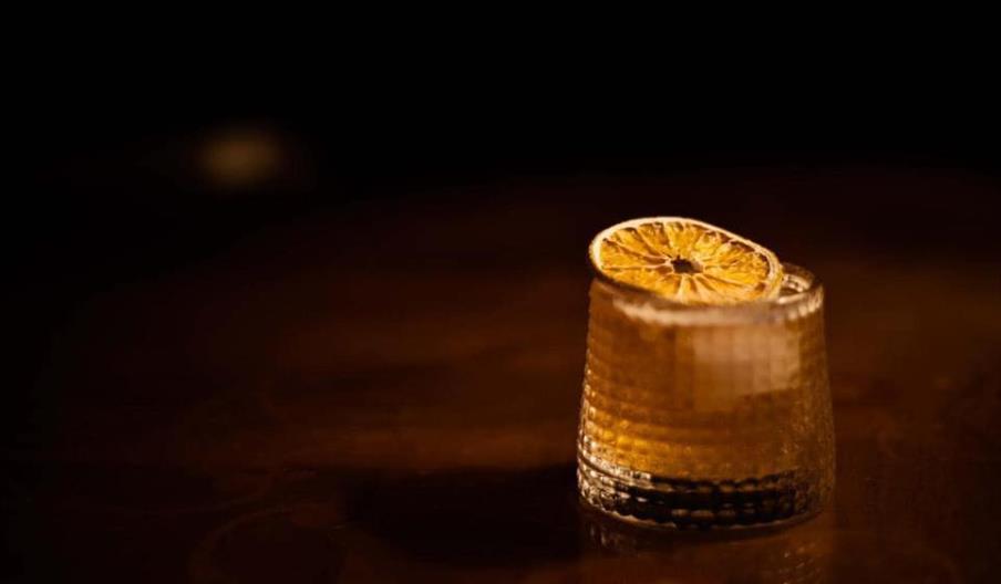 A textured glass filled with a dark beverage sits on a wooden surface, garnished with a dried orange slice, with a dark background.