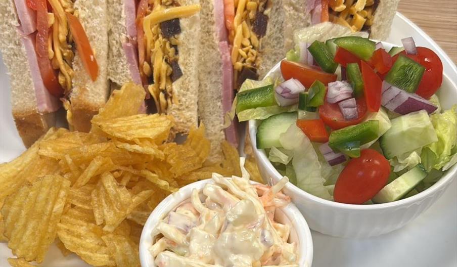 A plate with four club sandwich quarters filled with ham, cheese, bacon, and tomato, served with a side of ridged potato chips, a small bowl of creamy