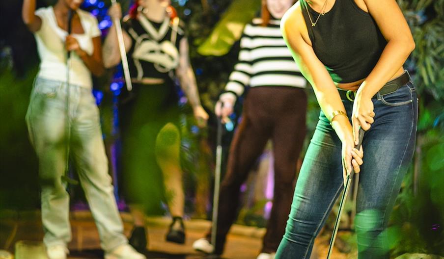 A person holding a golf club attempts to putt their ball into a hole as their friends