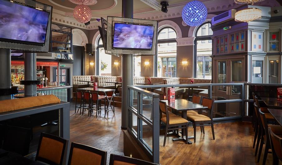 Inside view of  bar with tables, chairs and a bar in the corner and wall mounted TVs.